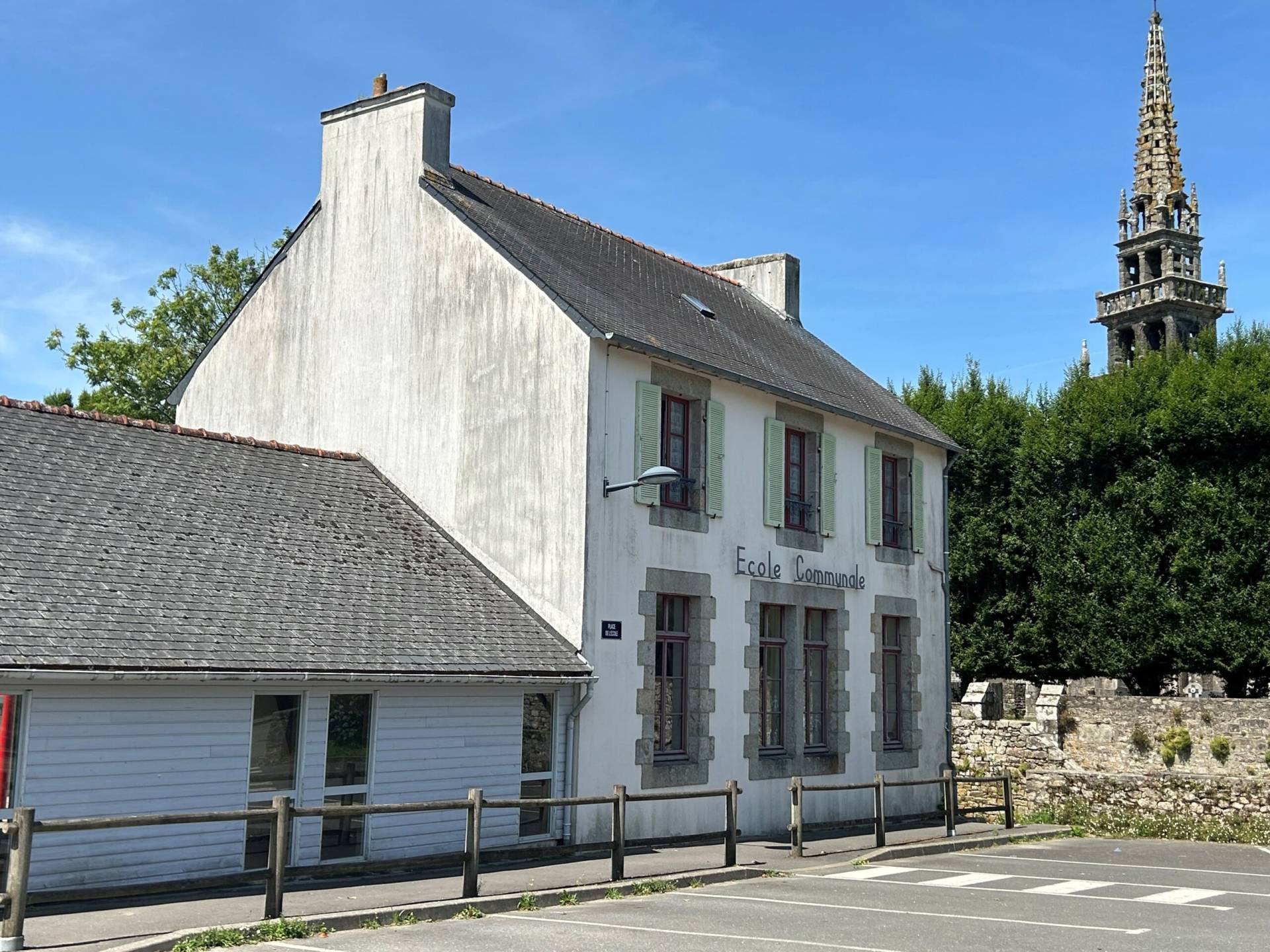 White school building with green shutters and stone-framed windows beneath a slate roof, blue sky above and a tall church spire in the background.
