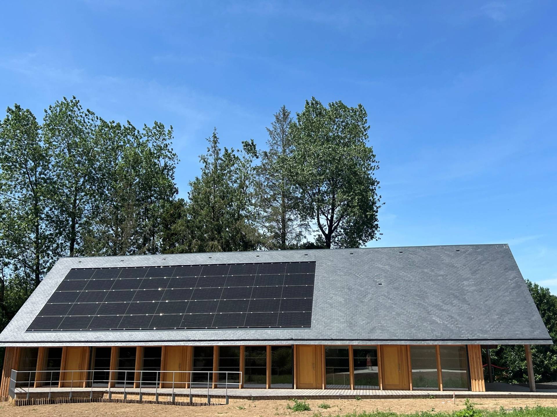 Building with a roof covered in solar panels, wooden entrance, and trees against a blue sky.