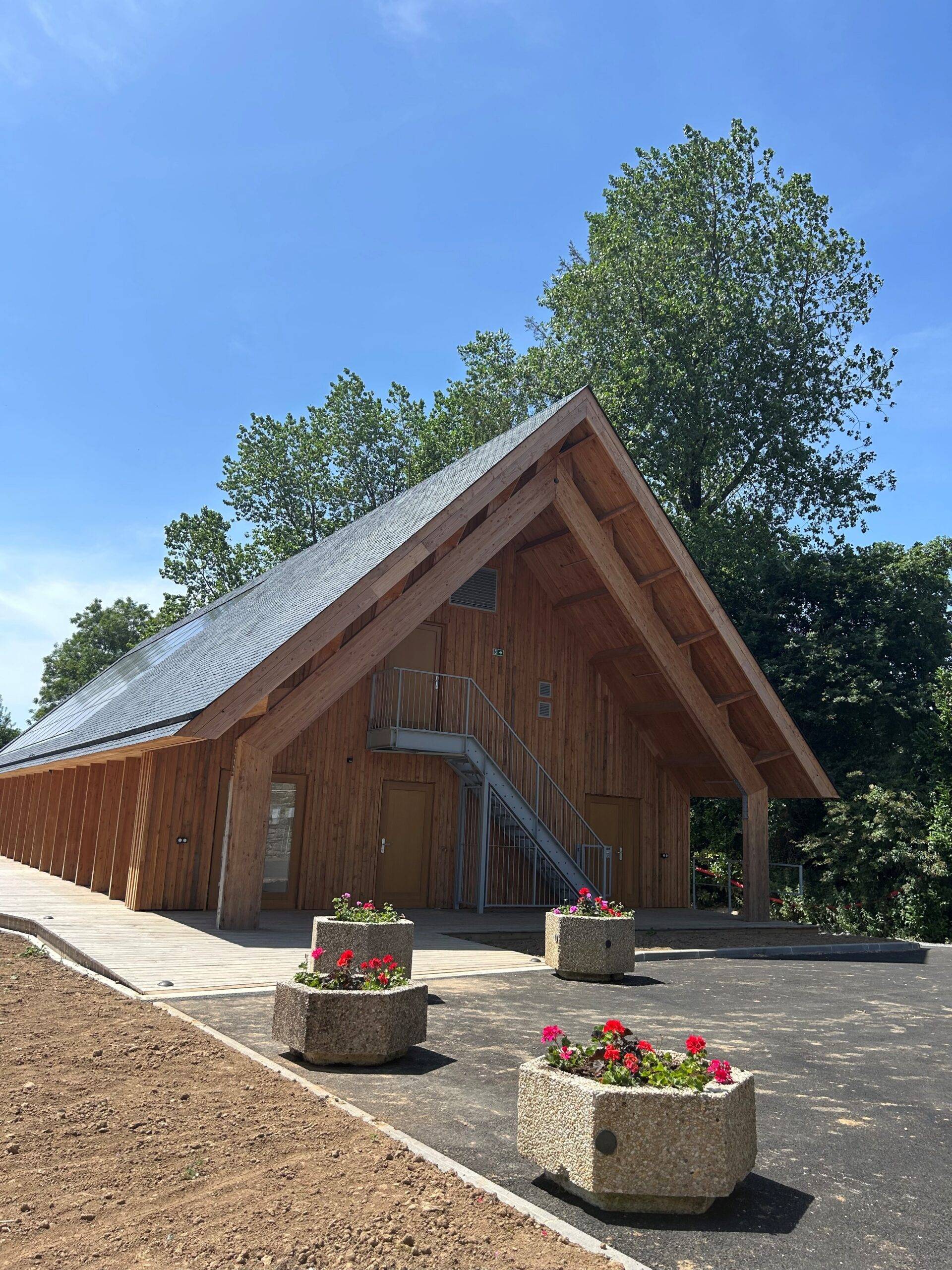 Wooden pavilion with a steep triangular roof, metal exterior stairs, and pink flowers in concrete planters against a bright blue sky.